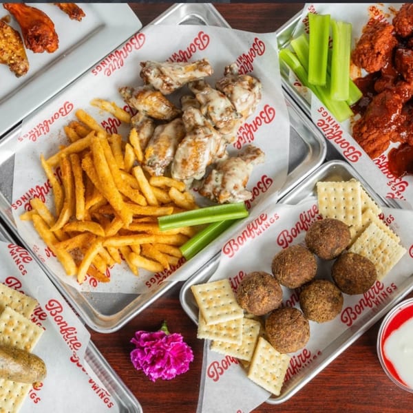 An overhead view of four metal trays of Bonfire Wings, fries, hush puppies, crackers, and celery.